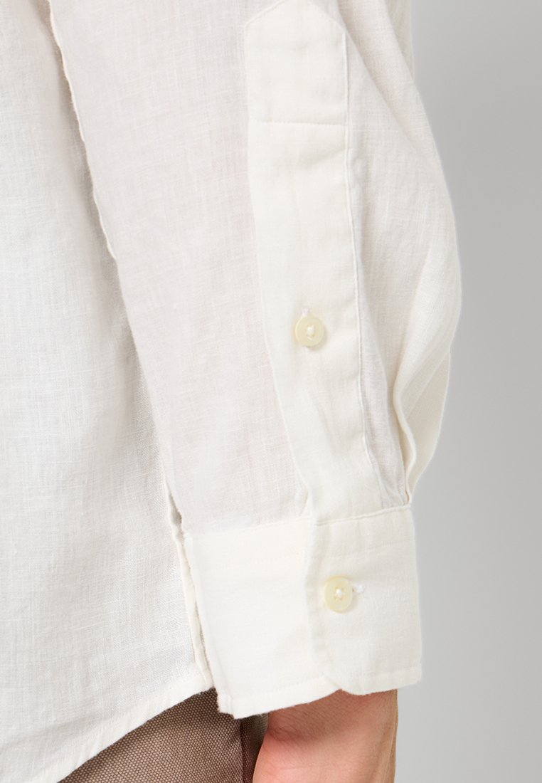 Chemise en lin couleur ivoire avec des poignets à bouton. Le tissu est léger et texturé, avec des fibres naturelles subtiles et des boutons doux.