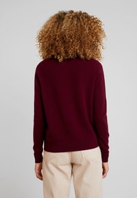 Maglione bordeaux con maniche lunghe, polsini e orlo a costine. Tessuto lavorato a maglia, scollatura rotonda e vestibilità rilassata. Dettagli di cucitura visibili.