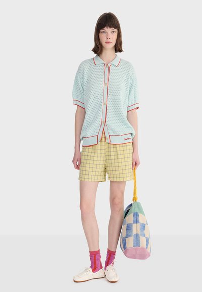 Femme portant un cardigan à manches courtes bleu clair avec des bordures rouges, un short à carreaux jaunes, des chaussettes roses, des chaussures blanches, tenant un sac à carreaux bleu et crème.