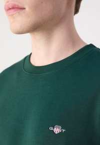 Jeune personne portant un sweat-shirt vert foncé à col rond avec un petit logo GANT brodé sur la poitrine.