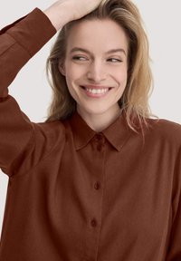 Donna con capelli biondi che sorride e guarda di lato, indossando una camicia marrone con bottoni, con una mano che tocca i capelli.