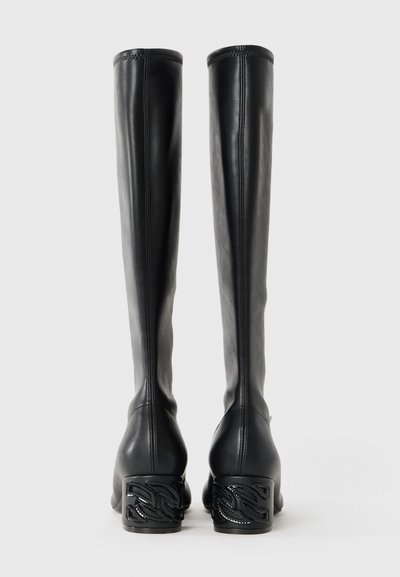 Par de botas de cuero negras hasta la rodilla con tacones bajos y un diseño de cadena en un fondo neutro.