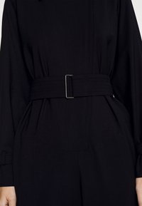 Mono negro con cintura ceñida, tejido texturizado y silueta estructurada con detalles plisados y resistente hebilla de plata.