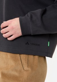 Maglietta a maniche lunghe grigio scuro con polsini a coste, dotata di un logo ricamato "Vaude" e un'etichetta verde sul lato in basso a sinistra.