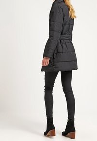 Musta toppatakki, jossa on vyötärövyö, huppu, tikattu pinta ja pitkät hihat. Päällä istuvien mustien farkkujen ja nilkkureiden kanssa.