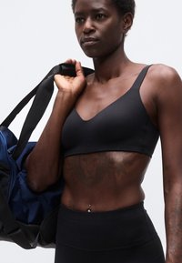 Αθλητικό άτομο με μαύρο αθλητικό σουτιέν και κολάν, κρατώντας μπλε και μαύρη τσάντα γυμναστηρίου στον ώμο, δείχνοντας το τατουάζ λιονταριού στον κορμό.