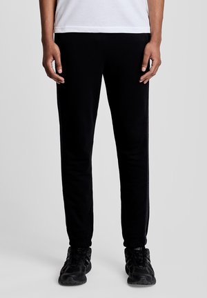 Persoon die zwarte taps toelopende joggingbroek en zwarte sneakers draagt, staand tegen een effen lichtgrijze achtergrond.