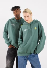 Felpe con cappuccio di un verde scuro, realizzate in un tessuto morbido, presentano una tasca frontale a marsupio e testo grafico; una felpa ha un emblema yin-yang giallo.