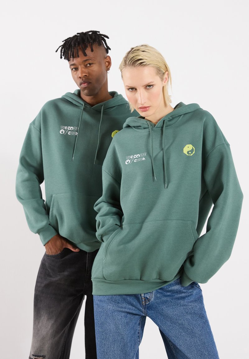 Sudaderas de color verde oscuro hechas de un tejido suave que cuentan con un bolsillo canguro frontal y texto gráfico; una sudadera tiene un emblema de yin-yang amarillo.