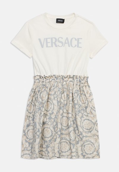 Λευκό φόρεμα T-shirt Versace με κοντά μανίκια, γκρι κείμενο λογότυπου στο πάνω μέρος και φούστα με μοτίβο λουλουδιών σε μπεζ και γκρι τόνους.