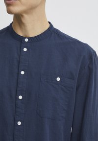 Marineblauw knoopshirt met een mandarin kraag, uitgerust met witte knopen en een rechthoekige borstzak. Zachte textuur.