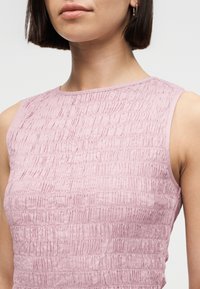 Γυναίκα που φοράει ροζ αμάνικο τοπ με υφή και οριζόντιο ριχτό μοτίβο, με κοντά ίσια σκούρα μαλλιά και μικρά σκουλαρίκια κρίκους.