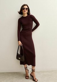 Abito a coste di un bordeaux scuro con maniche lunghe, silhouette aderente e lunghezza midi. Abbinato a sandali con cinturini marroni con tacco alto e una borsa marrone.