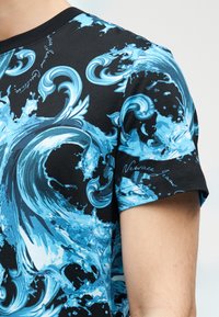Μαύρο κοντομάνικο πουκάμισο με σχέδια σε μπλε που απεικονίζουν κυματιστά νερά ωκεανού και πιτσιλιές νερού, φορεμένο στον άνω βραχίονα και τον ώμο ενός ατόμου.