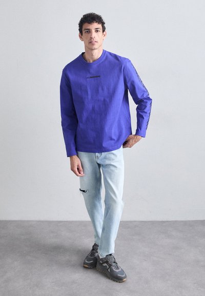 Chemise bleu à manches longues avec un col rond et un logo noir, associée à un jean léger bleu déchiré et des baskets foncées, se tenant sur un sol gris.
