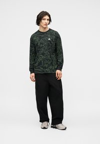 Νεαρός άνδρας που στέκεται φορώντας μαύρο μακρυμάνικο πουκάμισο με πράσινες αφηρημένες γραμμές, χαλαρό μαύρο παντελόνι cargo και μπεζ αθλητικά παπούτσια.