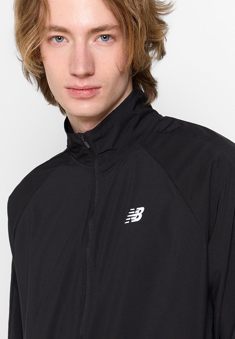 Zwarte jas met ritssluiting, gemaakt van lichtgewicht stof, met een hoge kraag en het witte New Balance-logo op de linkerkant van de borst. Glad oppervlakte.