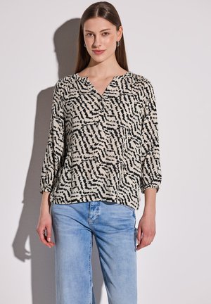 Leichte, cremefarbene Bluse mit schwarzem abstraktem Muster, V-Ausschnitt, Dreiviertelärmel, kombiniert mit hellblauen Jeans.
