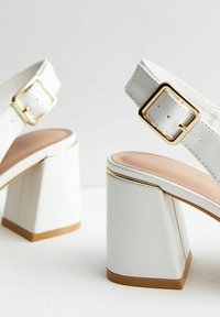 Bílé sandály na vysokém podpatku s blokovým podpatkem, vybavené zlatou sponou, hladkým povrchem a béžovou vložkou. Ozdobené zlatým rámem na podrážce.