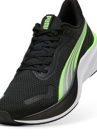 Zapatilla deportiva negra con material de malla, acentos en verde neón y plata, superficie texturizada, cordones y una suela blanca gruesa.