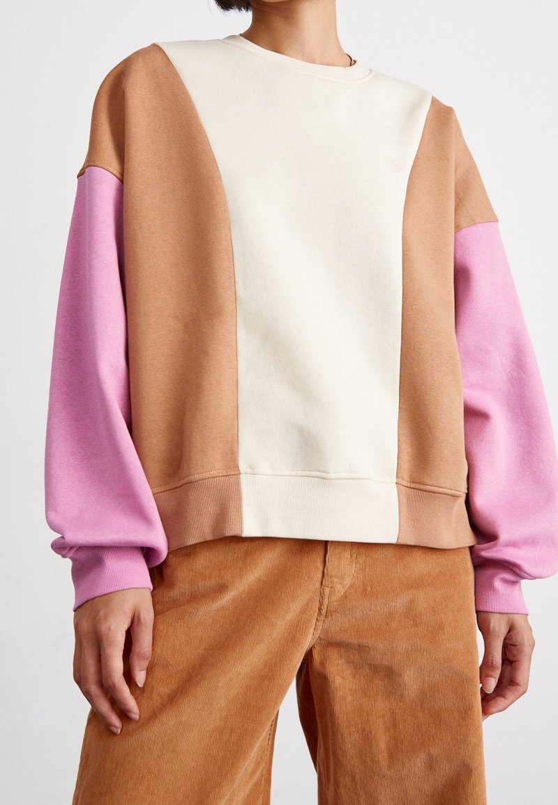 Personne portant un sweat-shirt color-block avec des sections beige, marron et rose, associé à un pantalon en velours côtelé marron, visible du cou aux hanches.