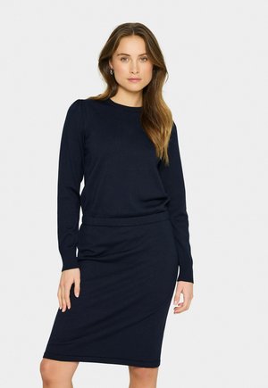 Laivansinis wool dress, jossa on pitkät hihat, ribbitekstuuri kauluksessa ja vartalonmyötäinen hame, joka ulottuu juuri polven ylle. Yksinkertainen, saumaton muotoilu.