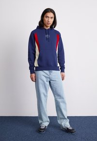 Haut à capuche marine avec panneaux latéraux rouges et crème, cordon de serrage réglable et poche frontale. Associé à un jean bleu clair et des baskets noires.