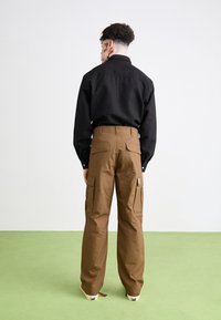 Braune Cargo-Hosen mit mehreren Taschen, aus robustem Stoff, kombiniert mit einem eng anliegenden schwarzen Hemd, von hinten gegen einen grünen Boden betrachtet.