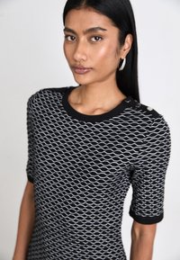 Black and white patterned top featuring a wavy design, short sleeves, ribbed neckline, and button accents on the shoulder.