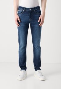 Vaqueros de denim azul con un ajuste regular, diseño de cinco bolsillos y costuras en contraste. Combinados con zapatillas blancas, mostrando un look casual.