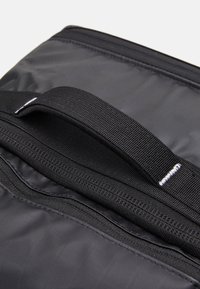 Svart nylonväska med en texturerad yta, med ett handtag på toppen och en synlig dragkedja. Sömmarna är i kontrasterande vitt.