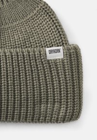 Olivazöld kötött sapka függőleges bordázott textúrával és egy fehér textilemke, amelyen a "DRYKORN" márkaneve szerepel.