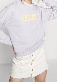 Liliowa bluza z żółtym logo LEVI'S, okrągły dekolt i ściągacze przy rękawach, połączona z białą spódnicą zapinaną na guziki. Miękka faktura materiału.