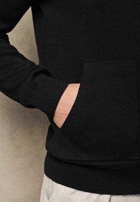 Černá mikina s texturovaným pleteným povrchem, se přední kapsou a žebrovanými manžetami. Detail ruky odpočívající v kapse.