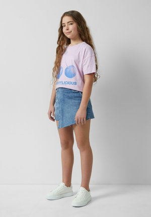 T-shirt rose clair avec un graphique de cerise bleue et le texte "CHERRYLICIOUS", associé à une jupe en denim et des baskets blanches.