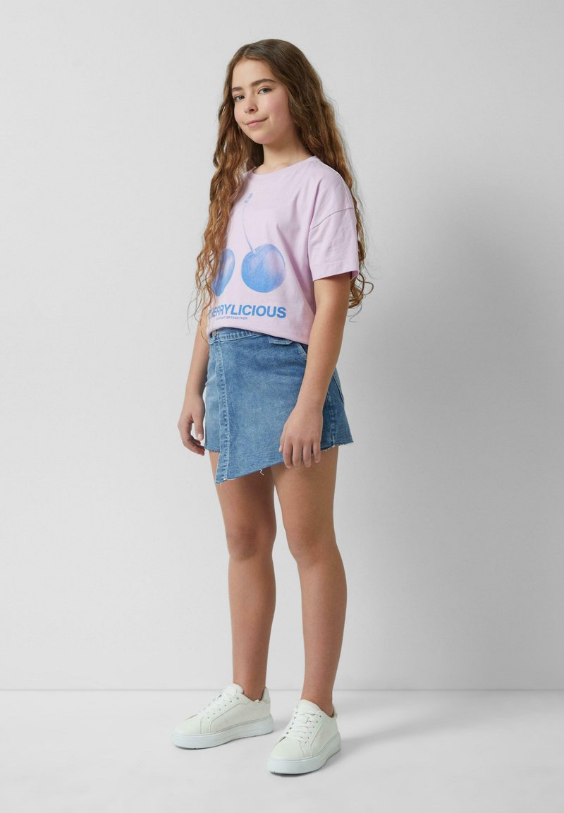 Helles rosa T-Shirt mit blauer Kirschgrafik und dem Text "CHERRYLICIOUS", kombiniert mit einem Jeansrock und weißen Turnschuhen.