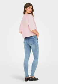 Kvinne står og ser over skulderen iført en lys rosa topp, lyseblå skinny jeans, brune loafers og hvite øreringer.