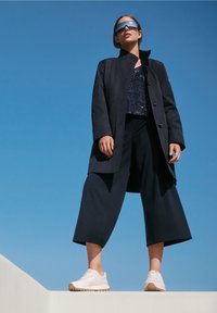 Un manteau noir sur un top à sequins, associé à un pantalon noir large et des baskets blanches. Comprend une semelle plate et des lunettes de soleil réfléchissantes.