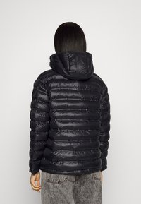 Černá péřová bunda s kapucí, s prošitými částmi, lesklým povrchem a elastickými manžetami, doplněná šedými kalhotami s vzorem.