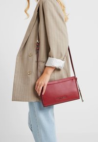 Kobieta w beżowej marynarce w paski i jasnoniebieskich dżinsach, trzymająca czerwoną torebkę na ramię Calvin Klein z regulowanym paskiem.