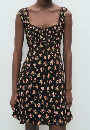 Femme portant une robe noire sans manches avec un imprimé floral rose et vert, corsage ajusté et encolure froncée.