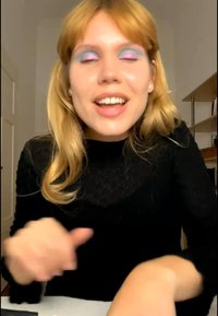Jonge vrouw met lichtbruin haar en kleurrijke blauw-roze oogschaduw glimlacht terwijl ze binnen zit, gekleed in een zwart topje. Handen zijn licht onscherp.