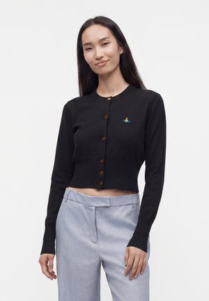 Ung kvinde iført sort knappet cropped cardigan med lille farverigt logo og lysegrå højtaljede bukser, stående på hvid baggrund.