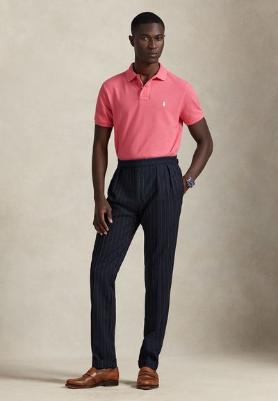 Uomo in piedi indossa una polo rosa a maniche corte, pantaloni blu navy a righe sottili, scarpe di pelle marroni e un orologio da polso su uno sfondo neutro.