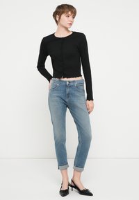 Črn rebrast dolg rokav krojen top z gumbom, v kombinaciji s svetlo modrimi pokončnimi kavbojkami in črnimi čevlji s konicami. Preprost, čist dizajn.