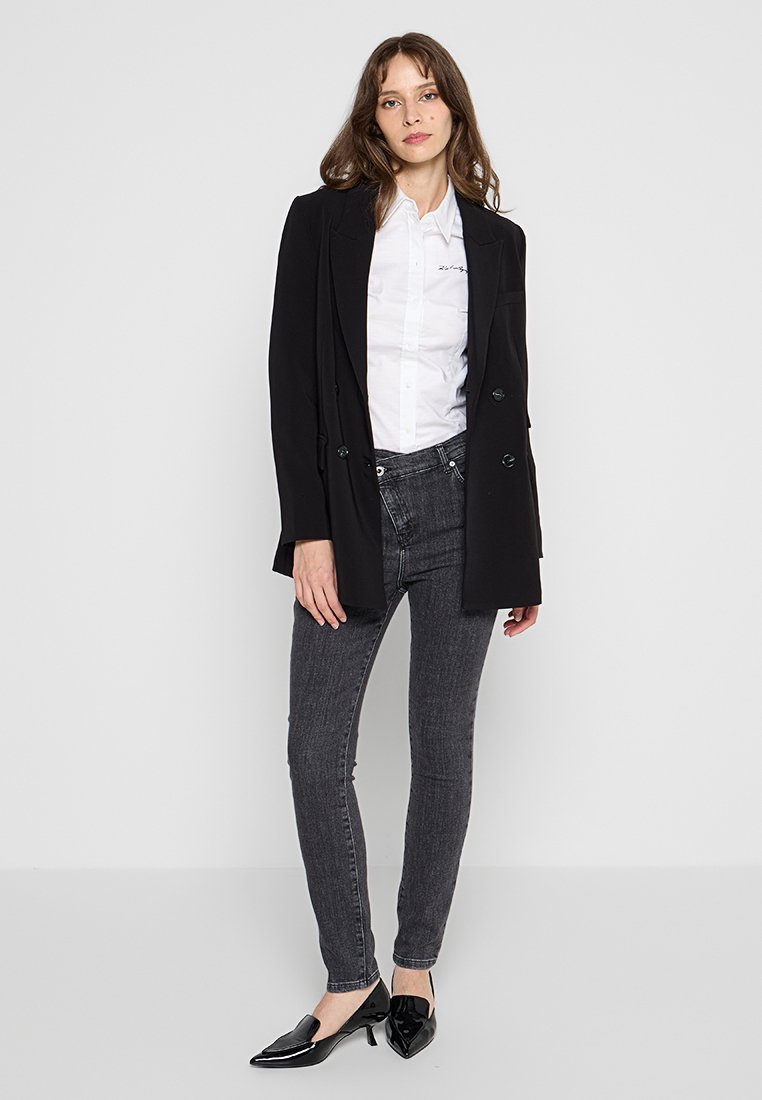 Blazer noir à double boutonnage sur une chemise blanche à boutons, associé à un jean skinny gris foncé et des chaussures noires à bout pointu. Textures lisses.