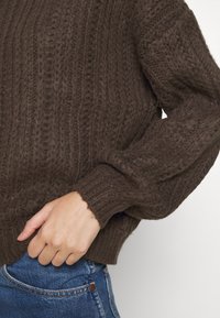 Brązowy sweter z dzianiny z teksturowanym żebrowanym wzorem, z długimi rękawami i luźnym krojem, zestawiony z niebieskimi dżinsami.