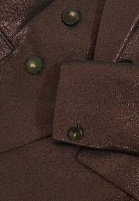 Hnědá metalická látka se strukturovaným povrchem, s velkými tmavými knoflíky a dvouřadým zapínáním. Detailní záběr na límec a manžety.