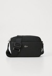 Fekete szövet keresztpántos táska állítható pánttal és ezüst Lacoste krokodil logóval az elején, egyszerű fehér háttér előtt.