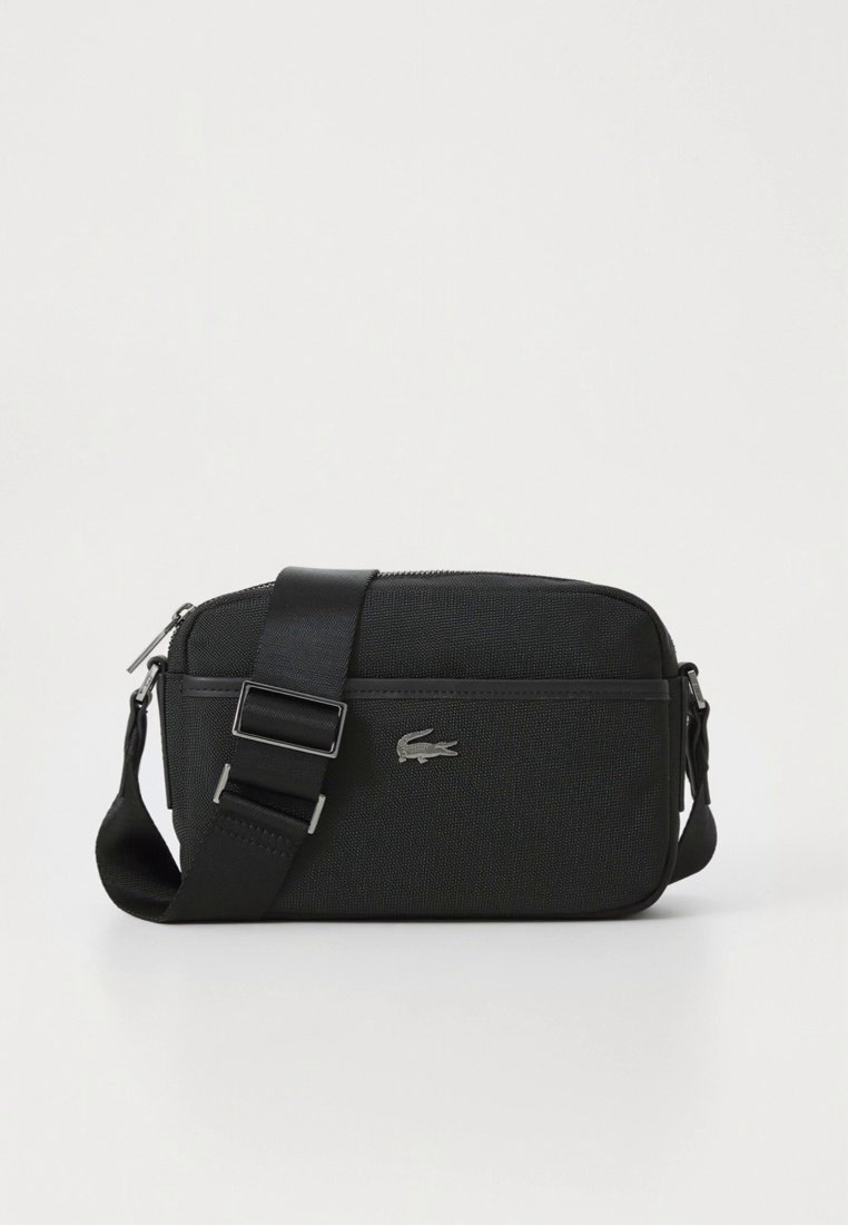 Sac bandoulière en tissu noir avec sangle ajustable et logo crocodile argenté Lacoste sur le devant, sur fond blanc uni.
